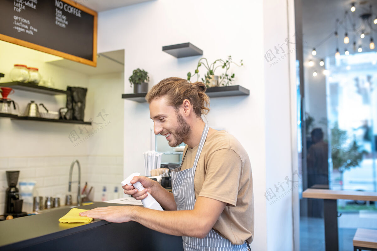 制服咖啡馆 清洁.long-一个留着胡子的年轻人在咖啡馆里打扫卫生 心情很好地擦拭着表面工作日围裙消毒剂