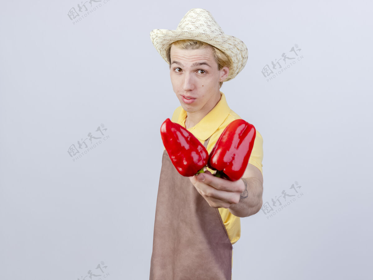 请高兴的年轻园丁男子穿着连体衣 戴着帽子 脸上露出微笑的红甜椒花园红色铃铛
