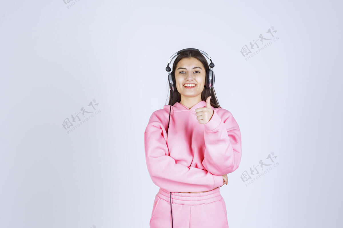 人类穿着粉色睡衣的女孩戴着耳机 展示着享受的标志高质量的照片休闲播放列表音乐