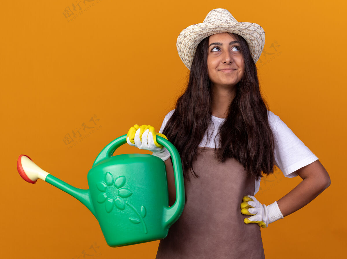 微笑穿着围裙 戴着夏帽 拿着喷壶的年轻园丁女孩站在橘色的墙上 脸上带着微笑抬起头来浇水花园年轻