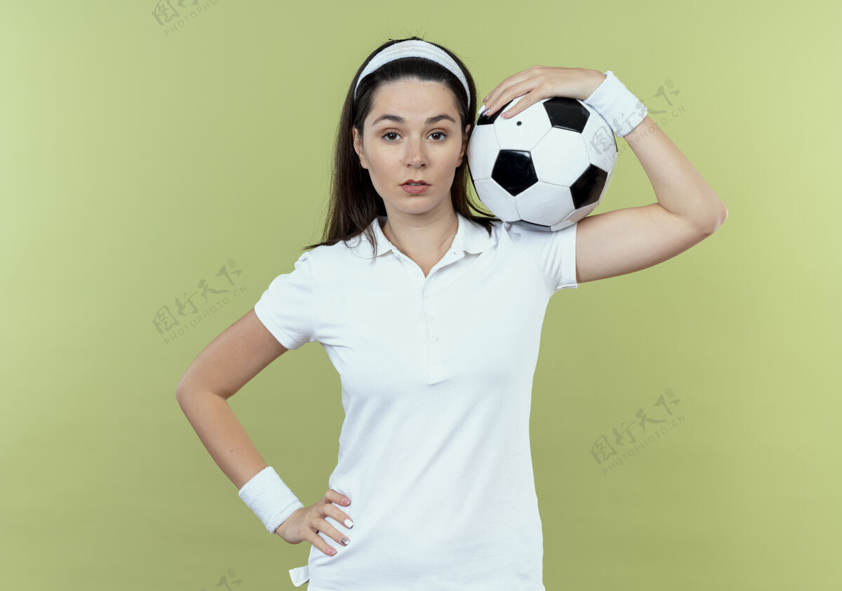 年轻戴着头巾的年轻健身女士肩上扛着足球 自信地站在轻盈的墙上举行表达轻