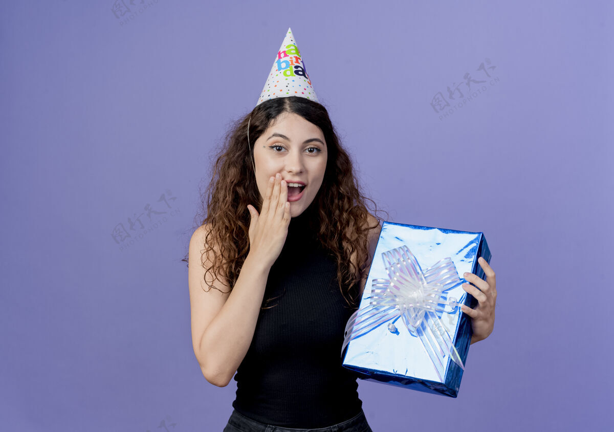 抱着年轻漂亮的女人 卷发 戴着节日帽 手里拿着生日礼盒 站在蓝色的墙上 看着令人惊叹的生日派对概念站着女人帽子