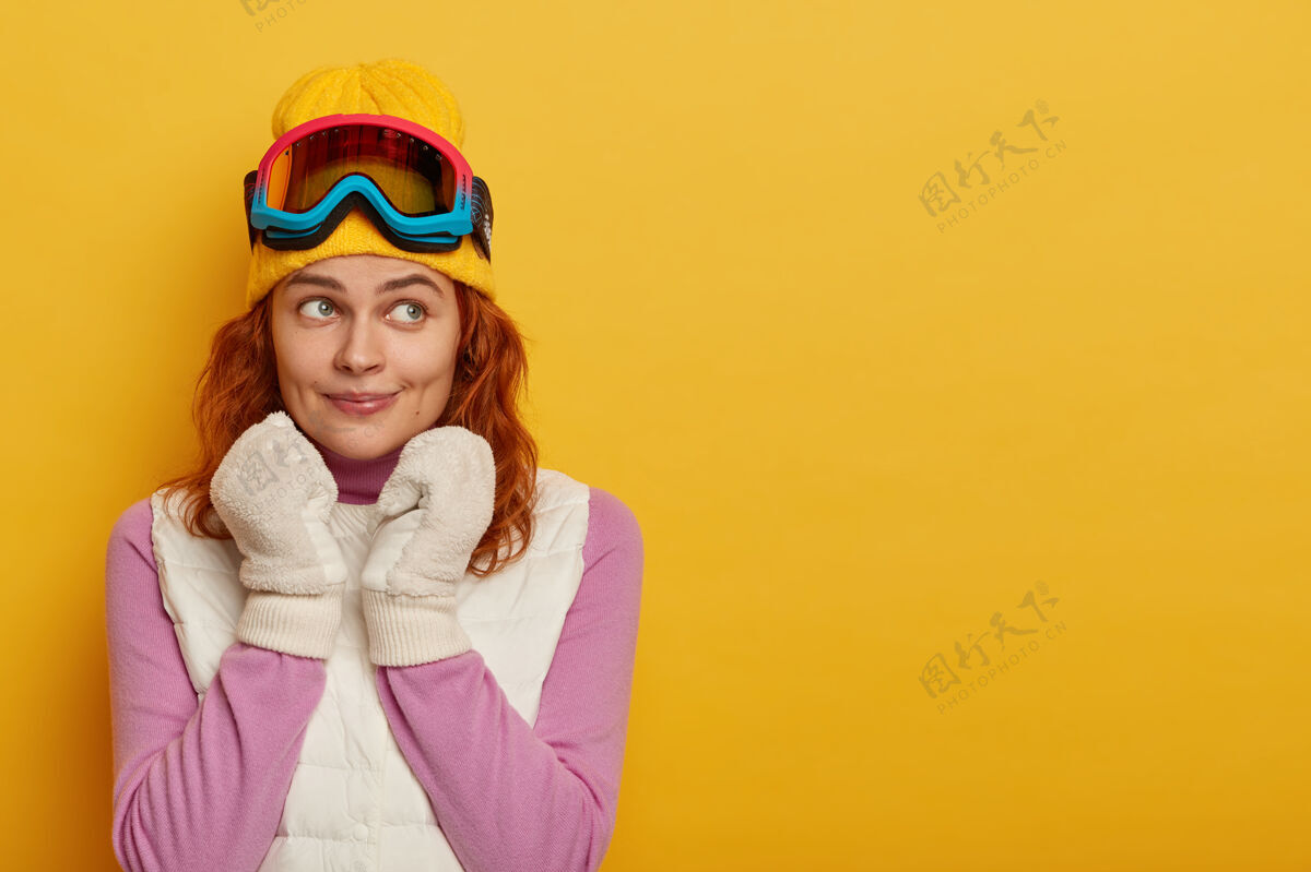 运动可爱的女游客红头发 头戴黄色帽子 头戴滑雪镜 戴白色软手套 穿紫色套头衫 穿白色马甲 下雪时积极休息 望向别处 在室内摆姿势成人室内年轻