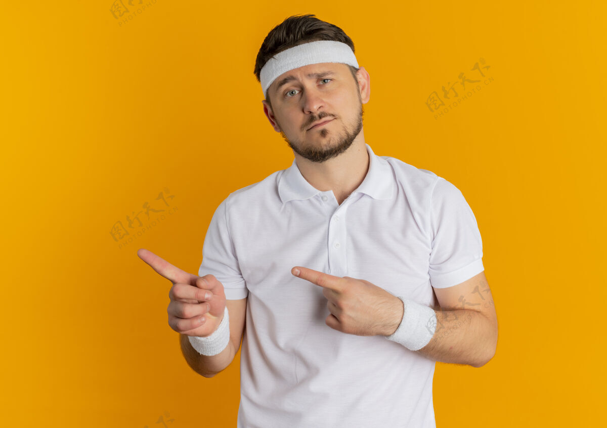 悲伤身穿白衬衫 头戴头巾的年轻健身男子面带悲伤的表情 手指指向站在橙色墙上的一侧男人头带表情