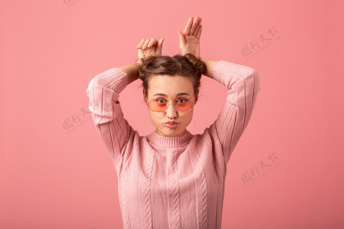 眼镜在粉红色的工作室背景下 穿着粉色毛衣 戴着太阳镜 表情滑稽的年轻漂亮可爱的女人的特写肖像 鬼混肖像女性表情