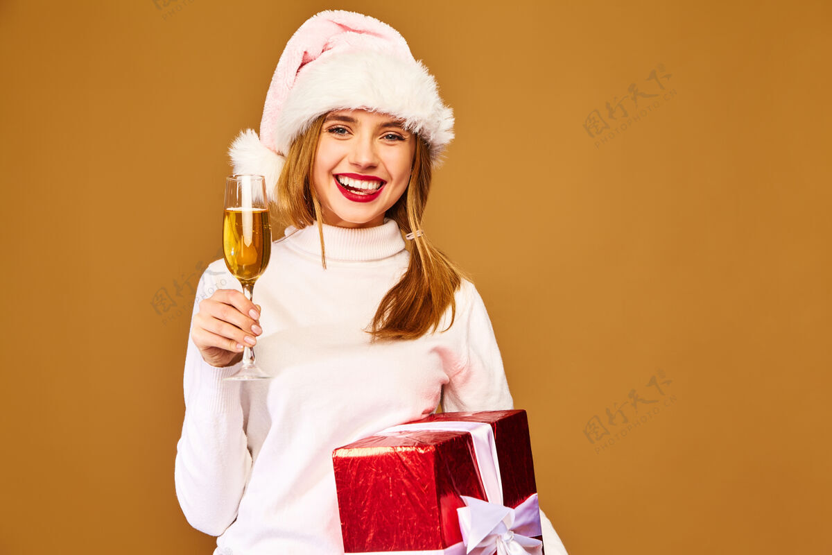 享受戴着圣诞帽和大礼盒的模特在金墙上喝香槟微笑帽子新年