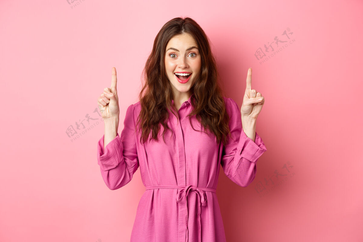 情绪迷人的快乐女士 手指向上 面带微笑 展示令人敬畏的广告 站在粉红色的墙上惊人休闲兴奋