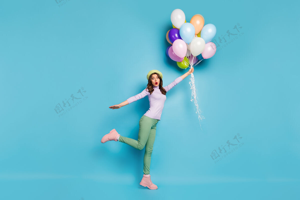 庆祝抓狂美女的全幅照片拿着许多气球惊天动地随风升起穿紫色套头衫贝雷帽绿色长裤靴子隔离蓝色墙壁空气惊喜冬季