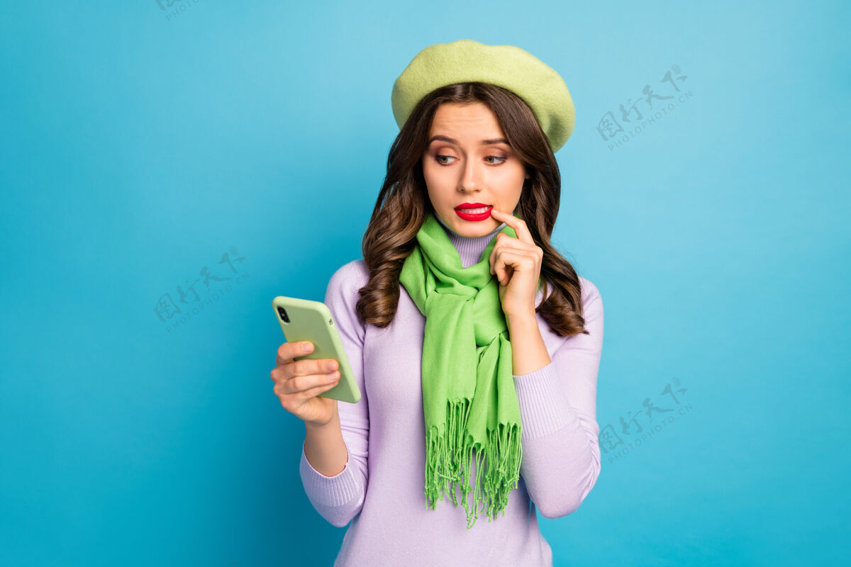 手机照片中美女手持电话双手咬手指有疑问新发帖文戴绿色贝雷帽紫色高领围巾隔离亮蓝色墙面高领毛衣不快乐阅读