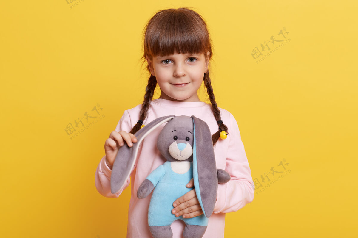 室内小孩抱着毛绒玩具动物看镜头 小女孩拿着她最喜欢的玩具玩 小女孩拿着毛绒兔子靠在黄色的墙上可爱温柔欢呼