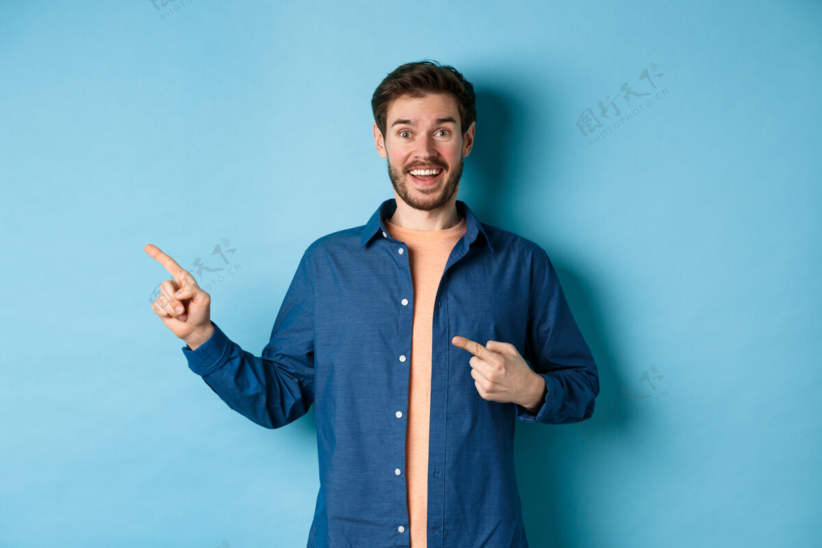 休闲兴奋和快乐的白人男子微笑着对着镜头 手指指着空旷的空间 展示标志 站在蓝色的背景上复制空间自信胡须成人
