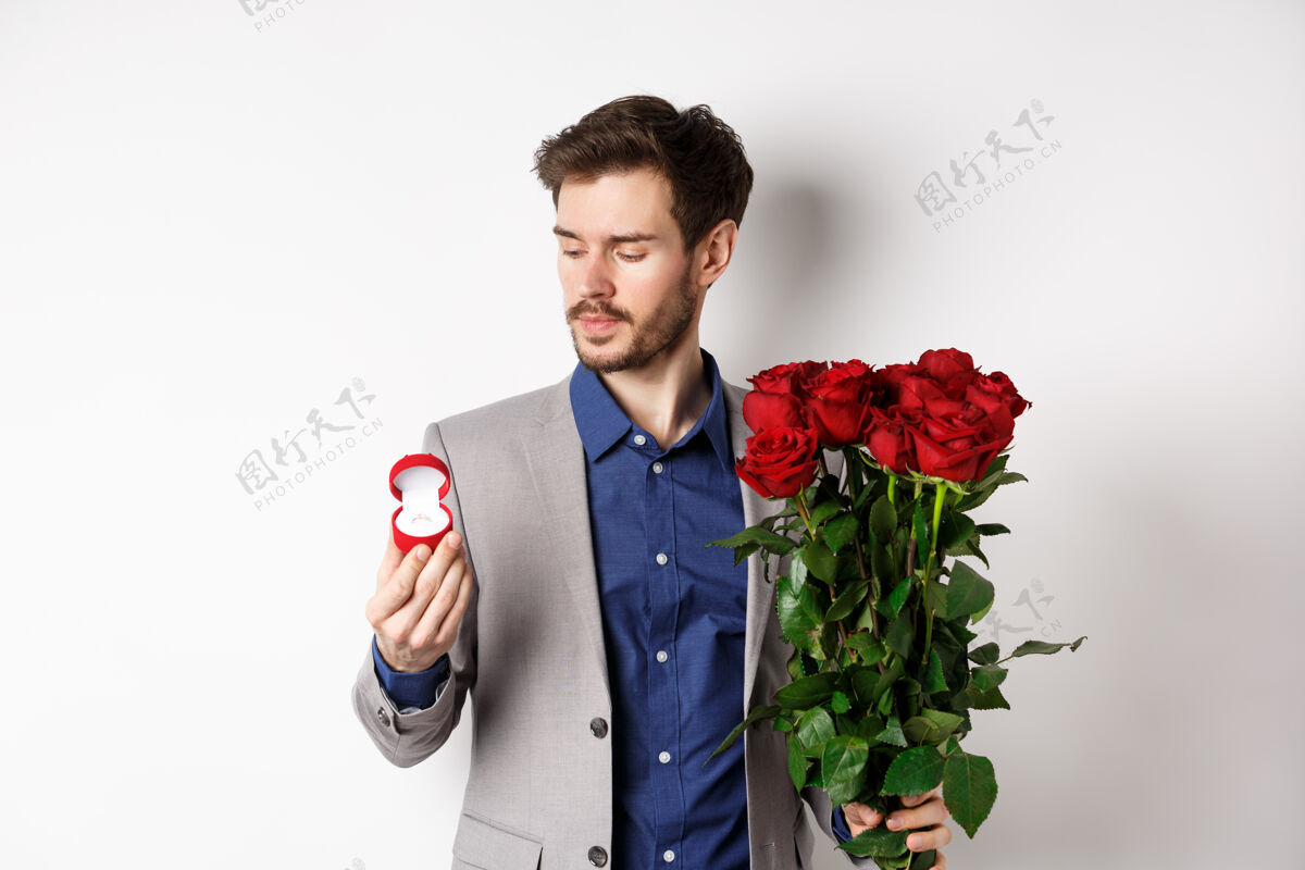 丈夫帅气的大胡子西装革履的男人看着订婚戒指 在情人节制造惊喜 站在白色背景下的红玫瑰套装男人感情