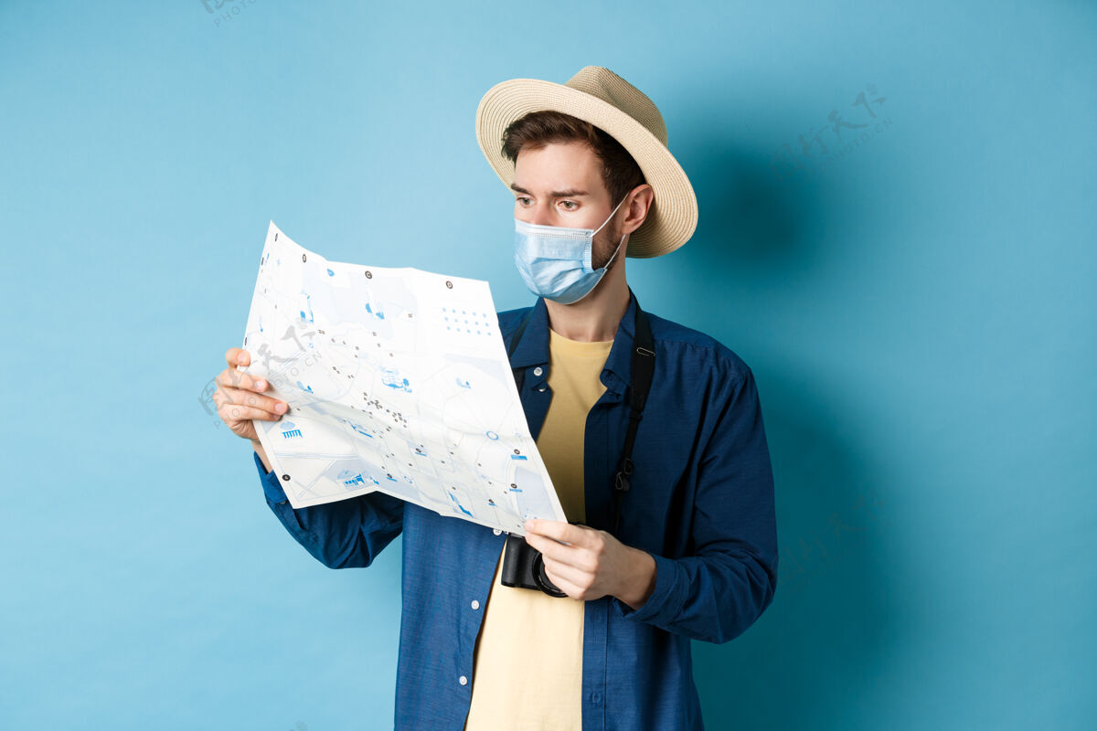 酷Covid-19 流行病和旅行概念戴着防暑帽 戴着口罩的帅哥 看着旅游地图 旅游度假 站在蓝色的背景上冠状病毒医学成人