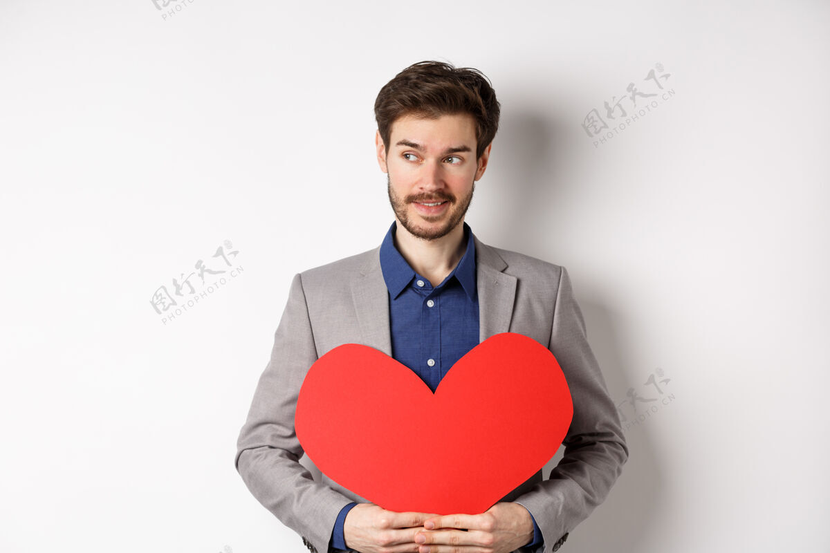 男人身着西装的帅哥左顾右盼 面带微笑 手拿红色心形剪纸 站在白色背景上为情人准备情人节惊喜。关系伴侣惊喜