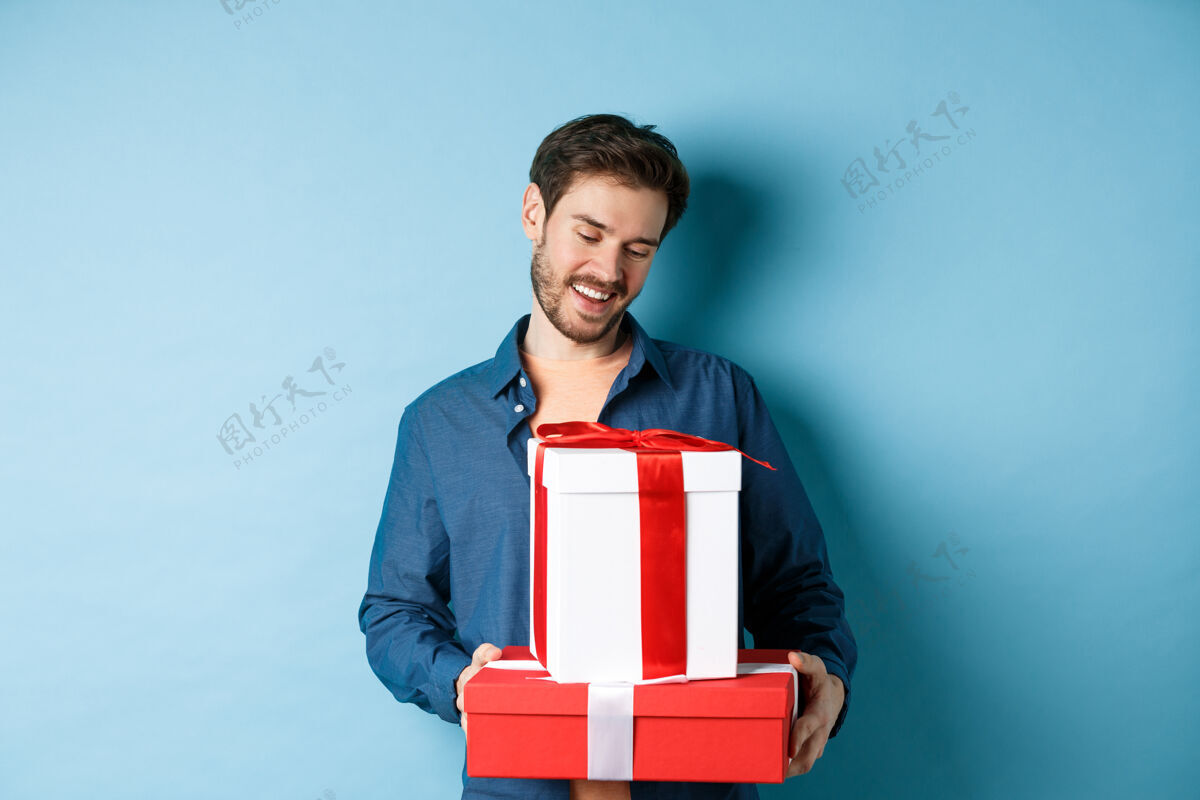 丈夫浪漫的年轻人 留着胡子 看着情人节的礼盒 给爱人送礼物 站在蓝色的背景上微笑男人温柔