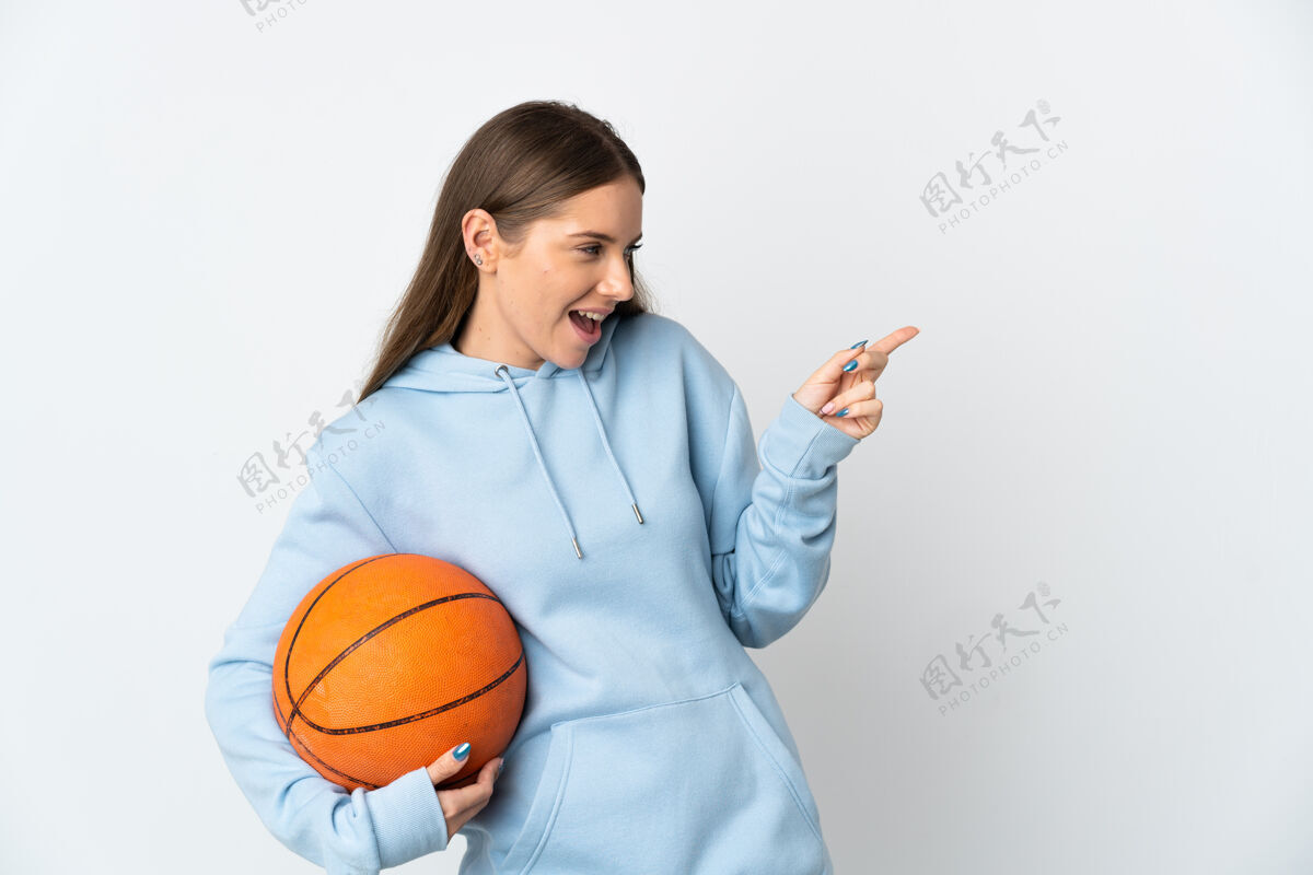 成人立陶宛年轻女子在白色背景上孤立地打篮球 手指指向侧面 展示产品运动员女孩壁板