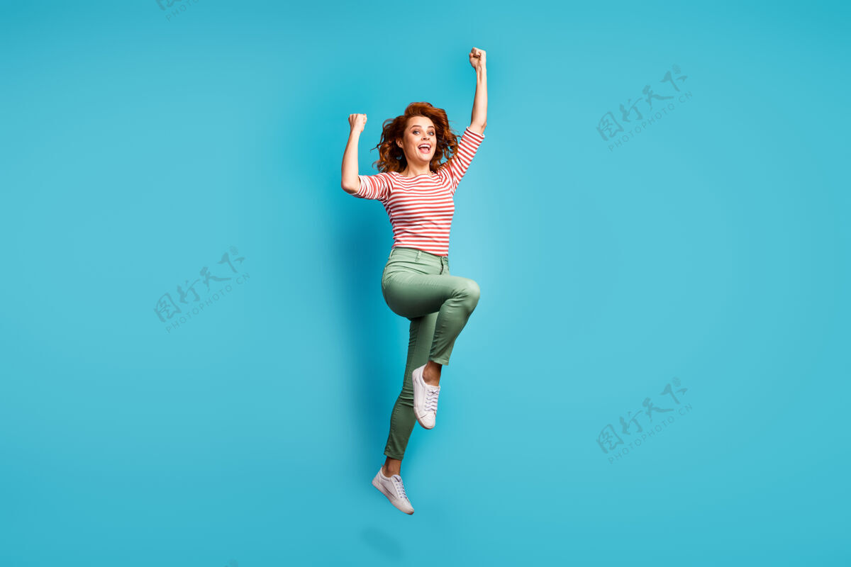运动搞笑女士跳高举起拳头赢得体育比赛冠军穿休闲红白衬衫绿裤子鞋孤立蓝色跳投飞行球迷