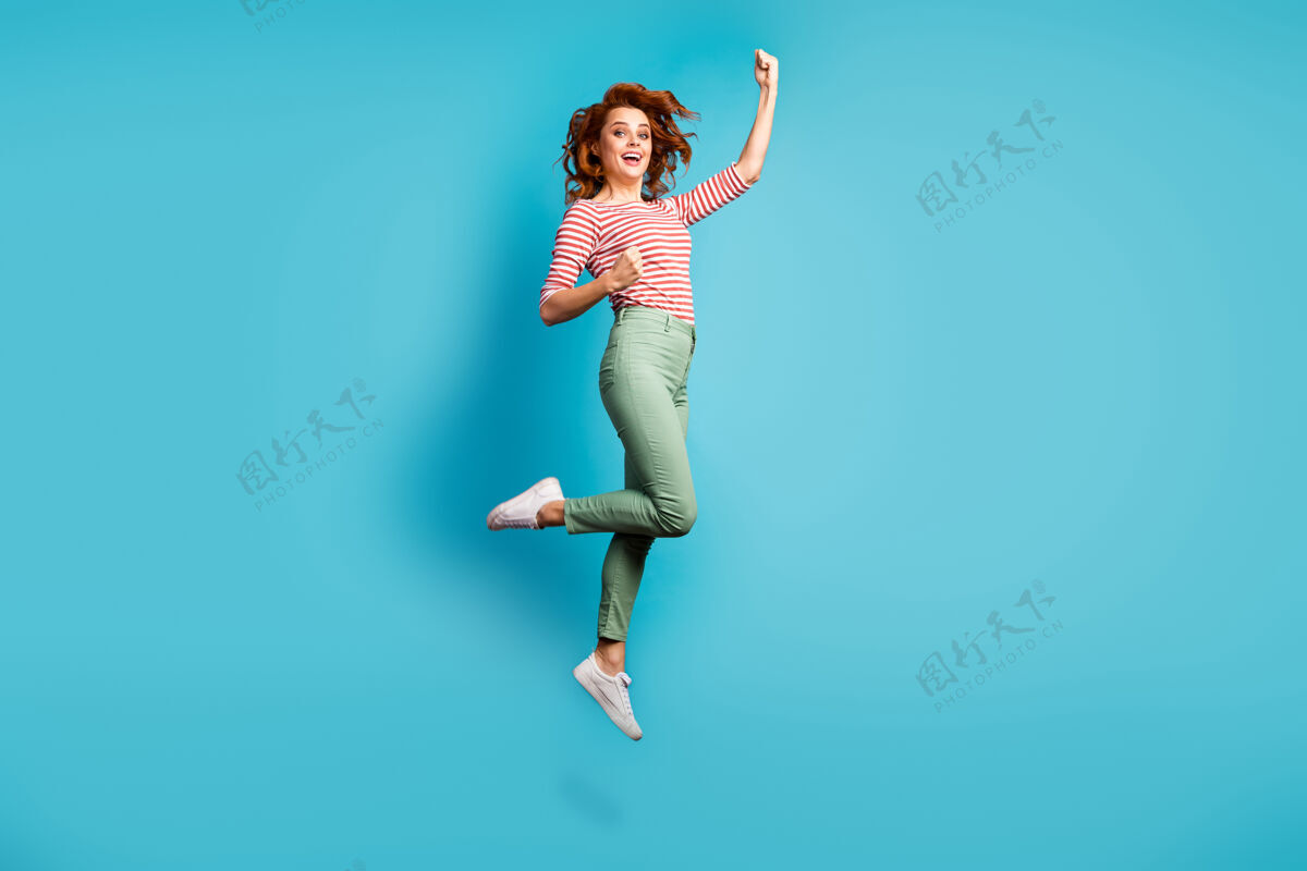惊讶搞笑女士跳高举起拳头赢得运动冠军庆祝穿休闲红白衬衫绿裤子鞋孤立蓝色时髦简介跳投