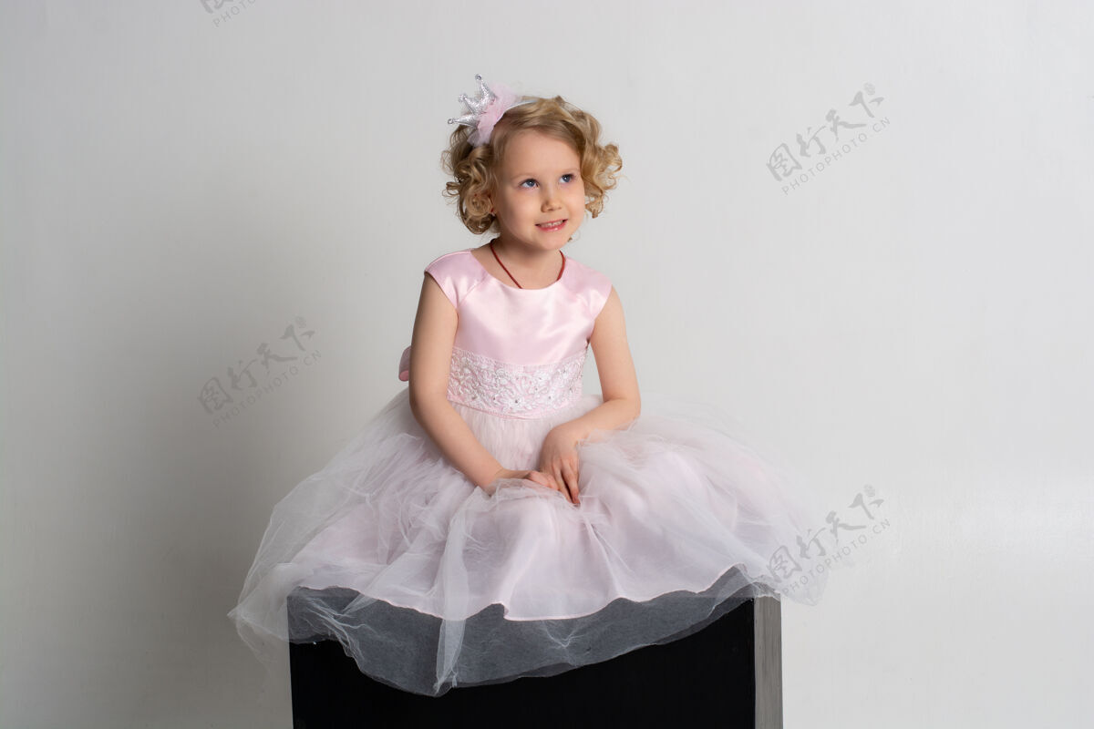 头发一个小女孩穿着粉红色的裙子 头戴皇冠 坐在白色背景的黑色立方体上年轻小美丽