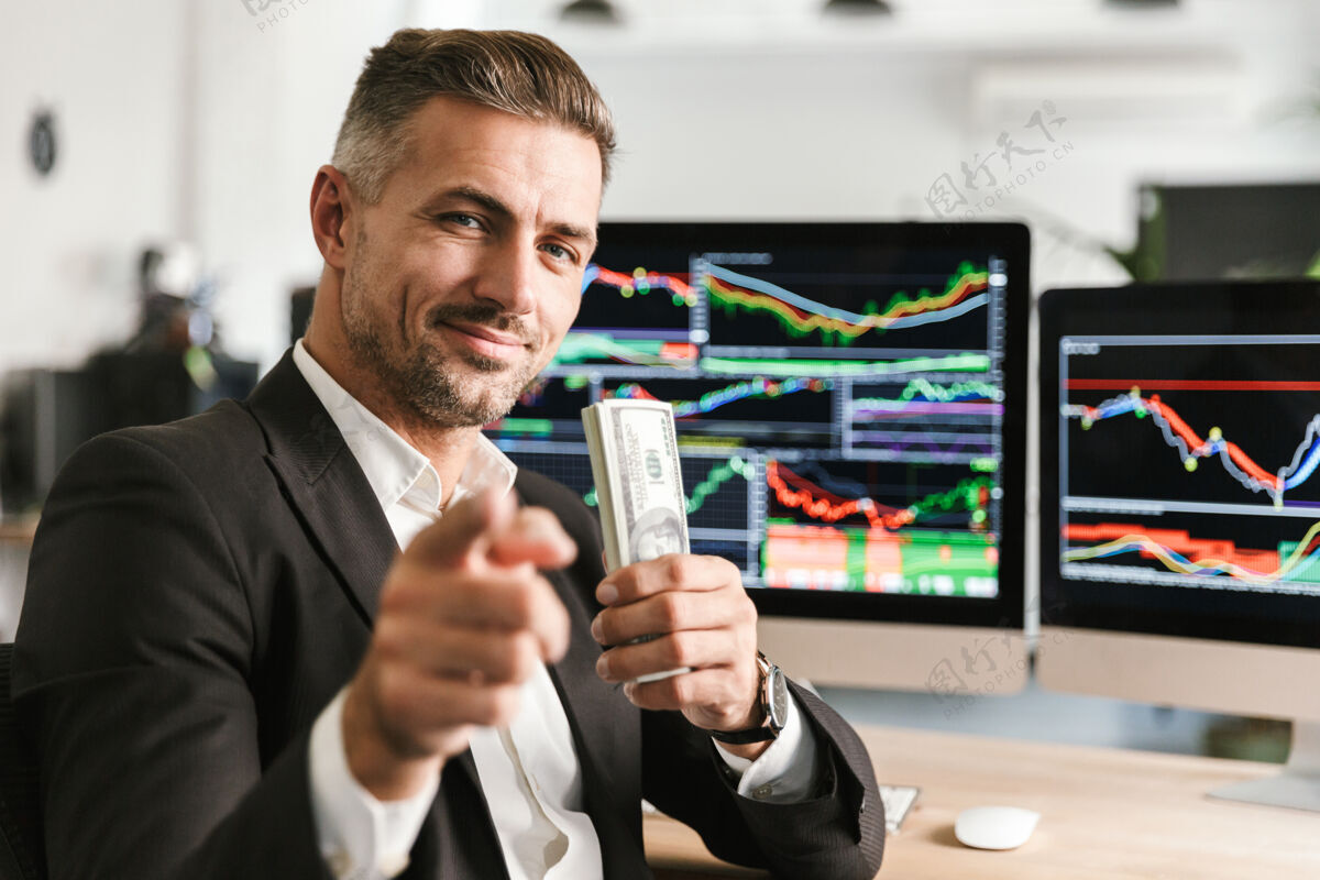 屏幕图为30多岁的白人商人穿着西装 手里拿着一包钱 在办公室工作 电脑上有图形和图表工作室内包装
