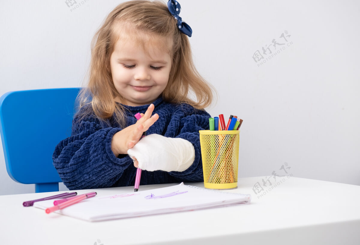 矫形坐在桌边用马克笔试着画画的小女孩手拿石膏手事故医学