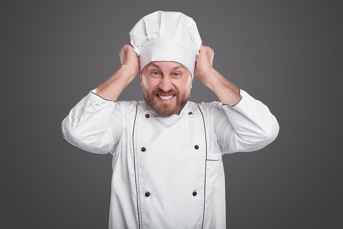麻烦愤怒的胡须男厨师看着相机 愤怒地拉着帽子 而在灰色背景下的餐厅工作愤怒恼怒错误