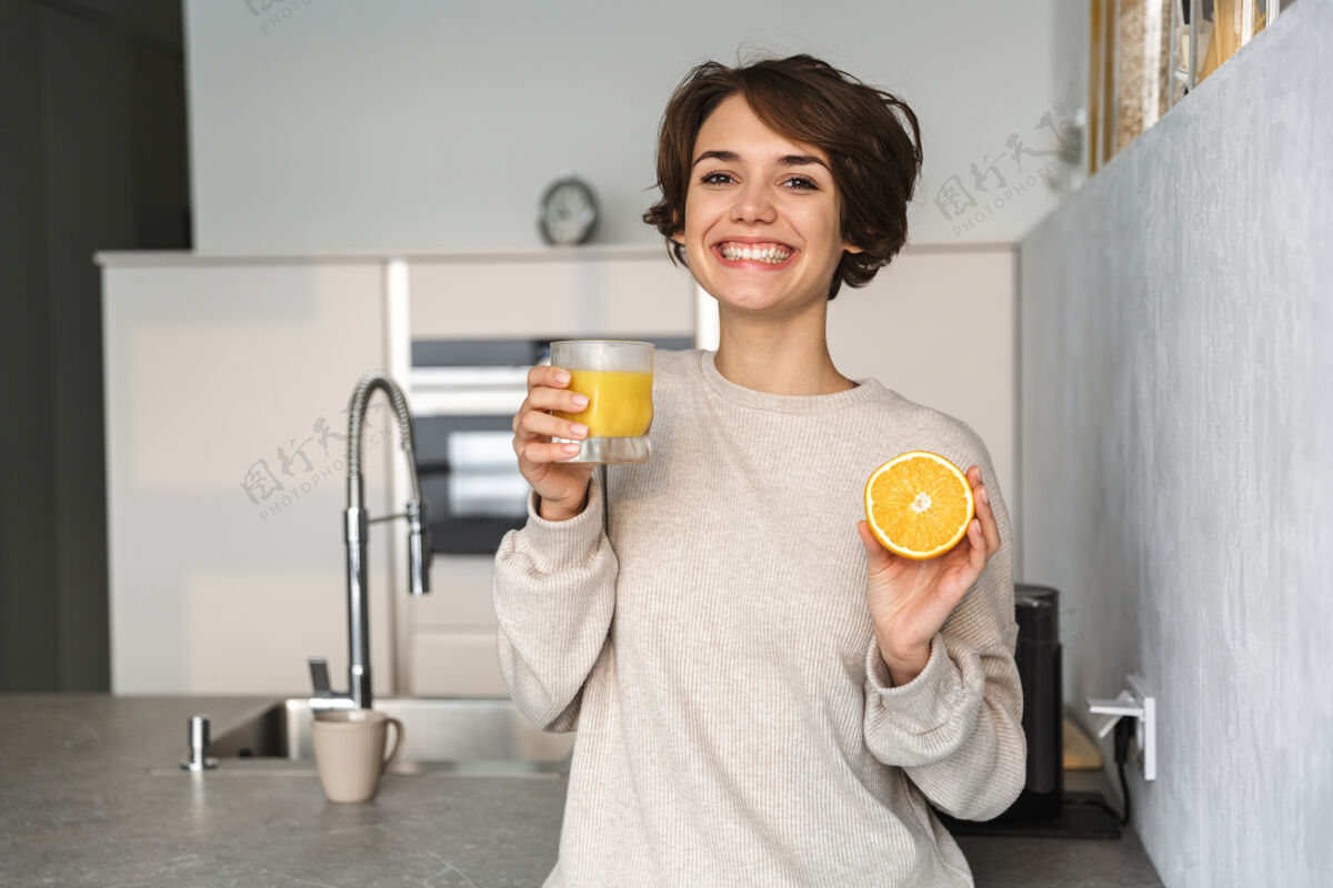制造站在厨房里 手里拿着橙汁和橙子水果的微笑的年轻女子成人能量控股