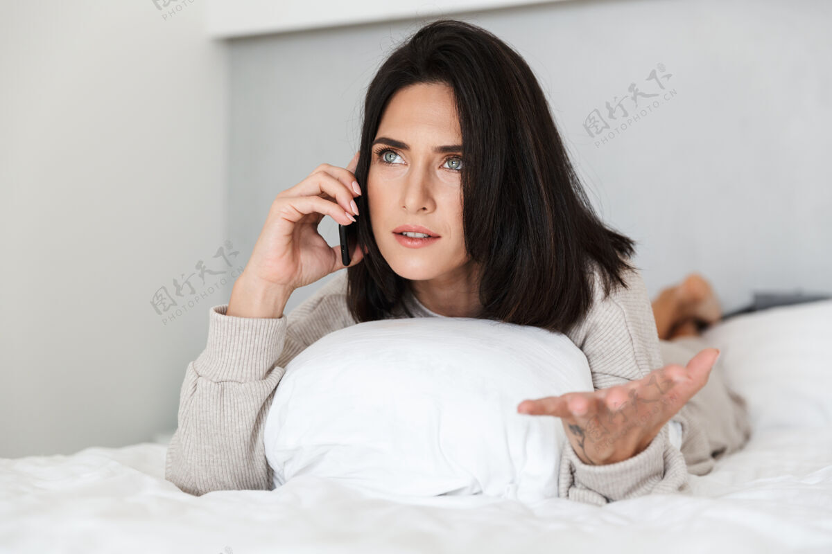 室内一张30多岁的漂亮女人在明亮的房间里躺在床上用手机的照片深褐色愉快舒适