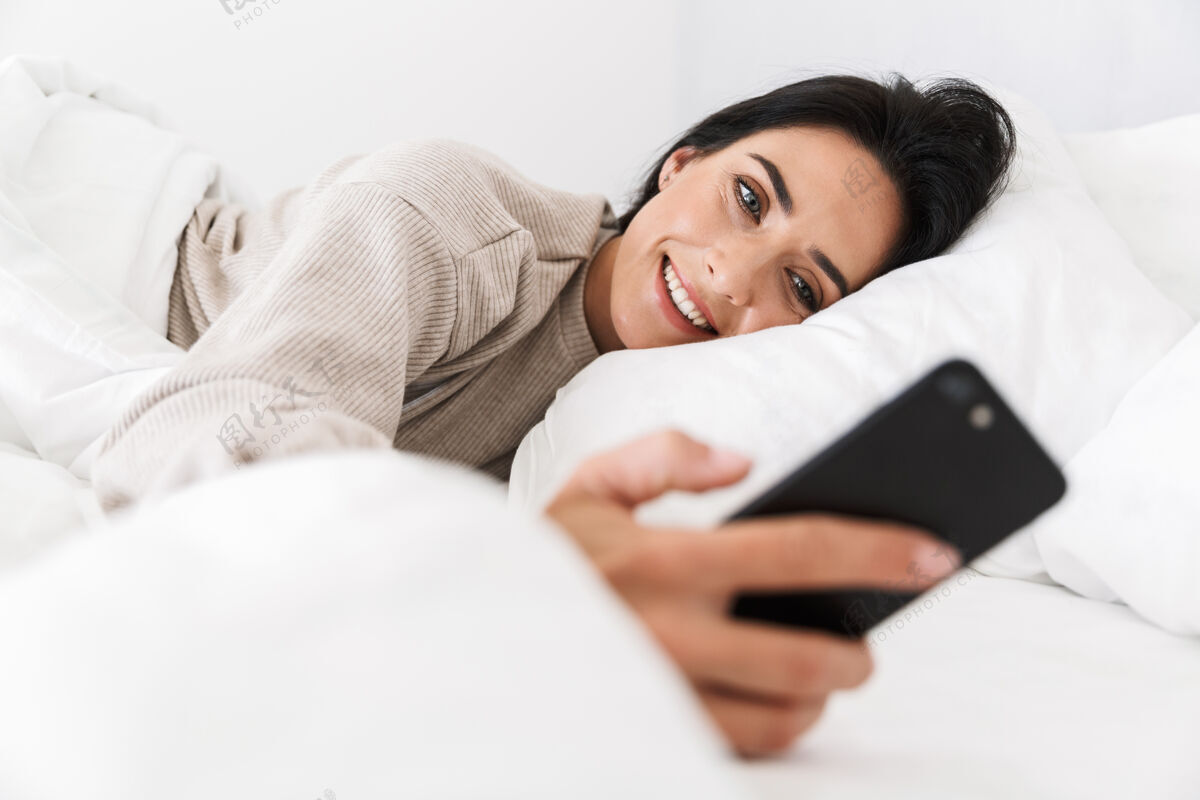 小玩意30多岁的美女在家里穿着白色亚麻布躺在床上用智能手机拍照公寓白种人愉快