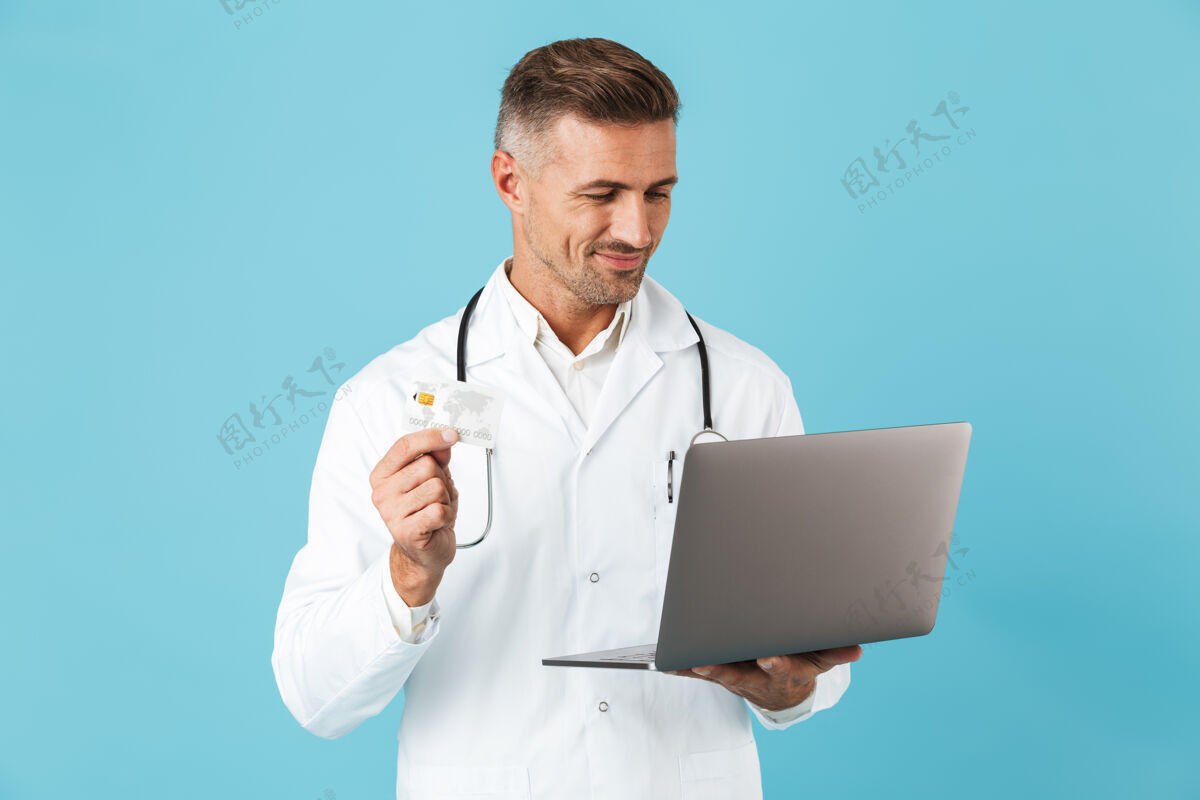 成人照片中的欧洲男子身穿白色医用外套 手持笔记本电脑和信用卡 手持听诊器 孤立地站在蓝色的墙上男人积极医生