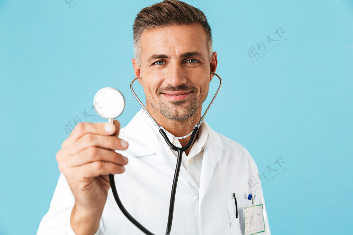 成熟微笑的医生的画像穿着白色制服手持听诊器 孤立地站在蓝色的墙上男人中年肖像