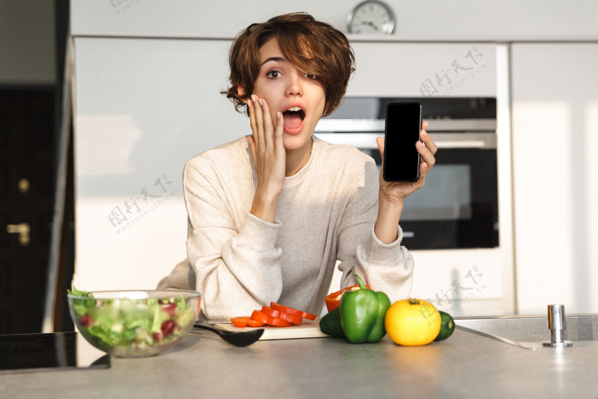 手机在厨房的桌子旁做饭时 一位黑发女士惊讶地展示了一个空白的智能手机屏幕看黑发震惊