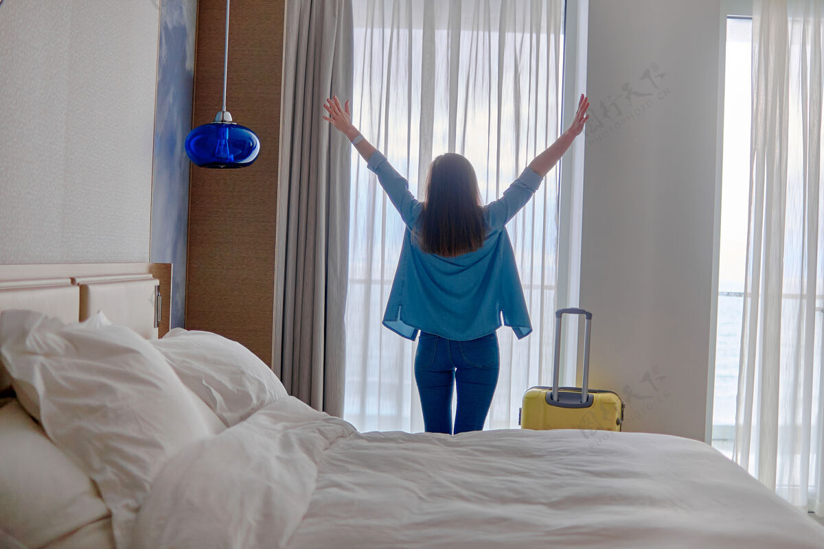 休闲一位快乐的旅客女士 举手提着手提箱 站在窗边 在登记入住后从酒店房间眺望风景享受室内客人