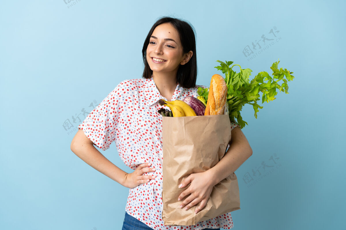 女性一个年轻的女人拿着一个杂货店购物袋摆姿势 双臂放在臀部 微笑着营养外卖购物者