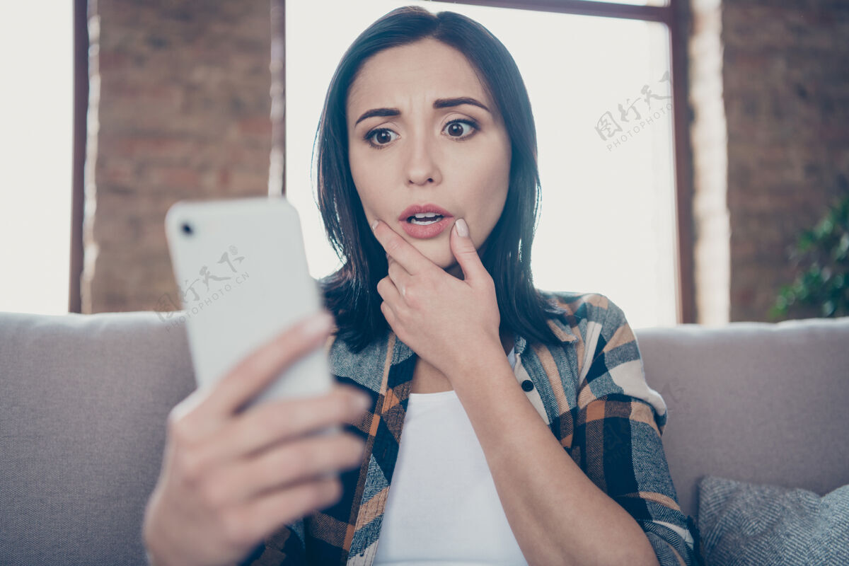 应用程序照片中美女手持电话不相信眼睛阅读新博文负面评论坐在舒适的沙发上穿着休闲服公寓室内房间房子不快乐