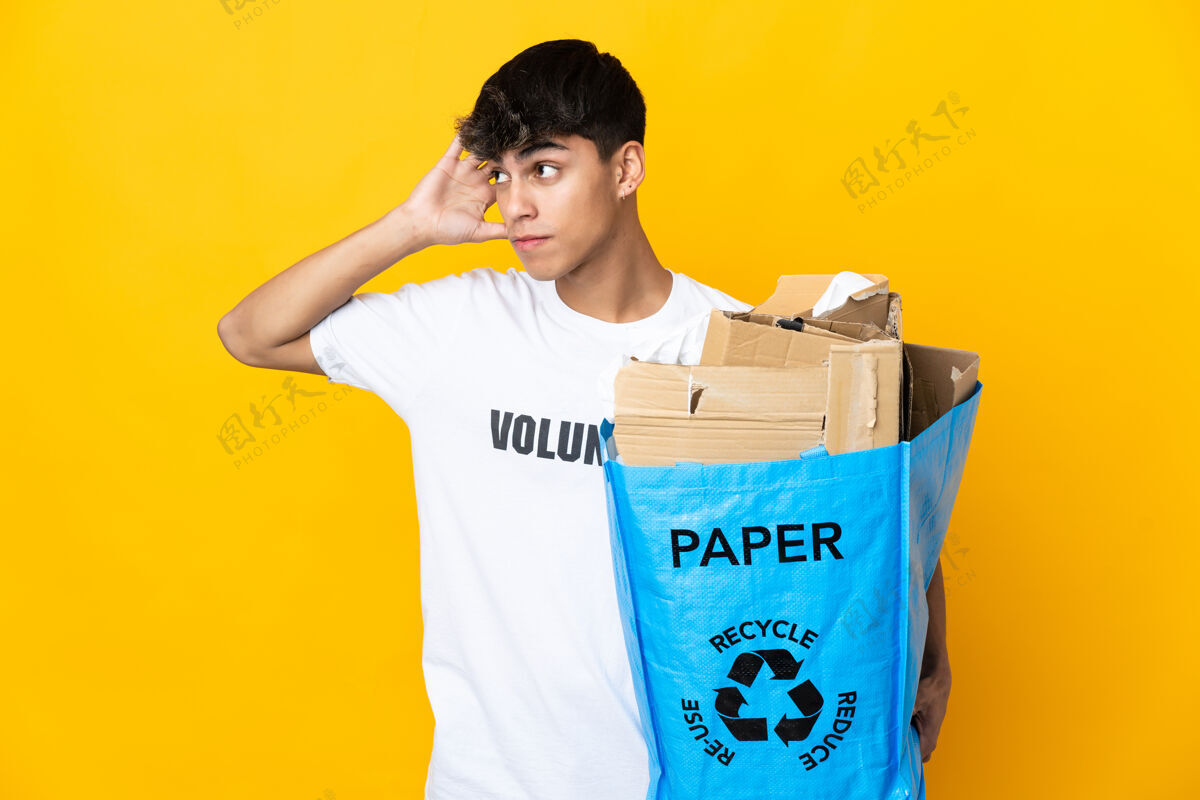 责任一个男人拿着一个装满纸的回收袋在孤立的黄色背景上进行回收 手放在耳朵上听着什么生态秘密年轻