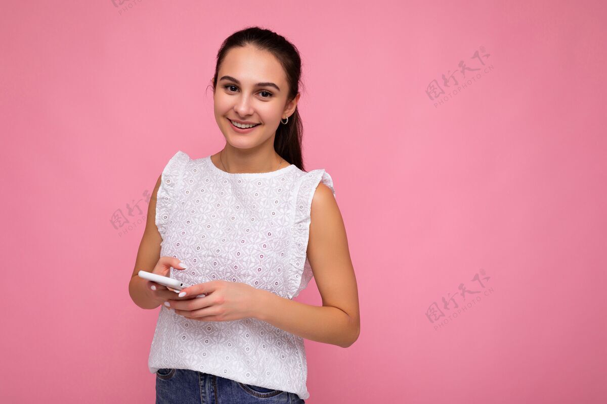 互联网漂亮的正面女孩穿着白色上衣隔着粉色背景墙用手机上网上网看照相机.副本空间手机成人浏览