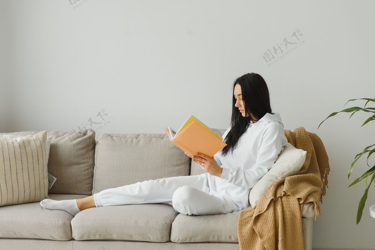 休闲漂亮的年轻女子躺在沙发上享受着在家看书的乐趣 穿着休闲装微笑着沙发坐着抱着