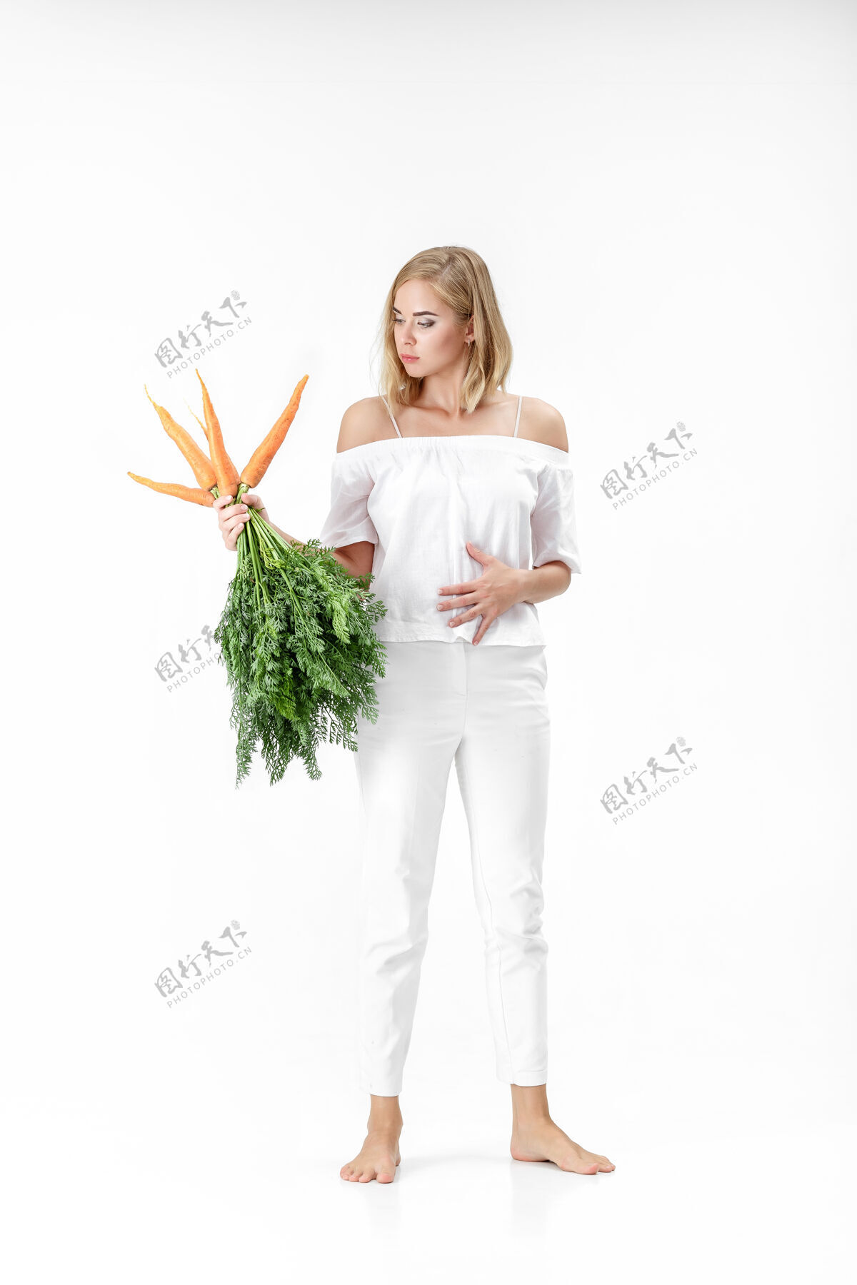 生一个穿着白衬衫的金发美女 手里拿着一根新鲜的胡萝卜 上面有绿叶背景.健康还有节食背景橙色脸