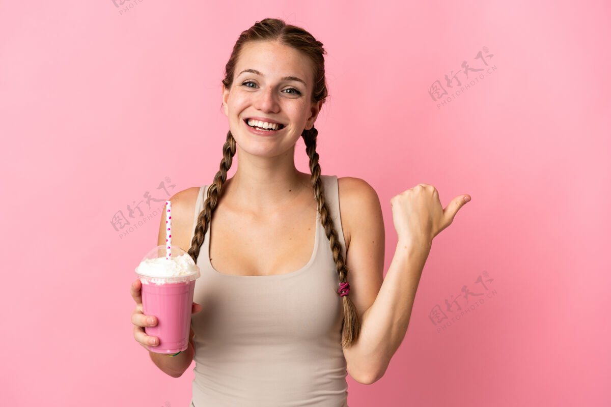 信心年轻女子用草莓奶昔隔离在粉色背景上 指着侧面呈现产品食物右女性