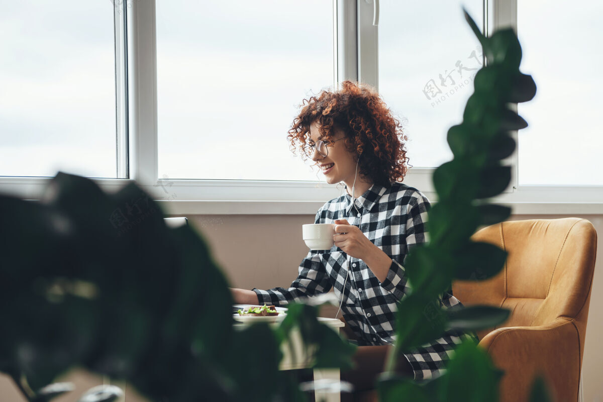 年轻人微笑的年轻女商人卷发 戴着眼镜 一边喝着茶 一边在笔记本电脑前吃着东西饮料生活方式浏览