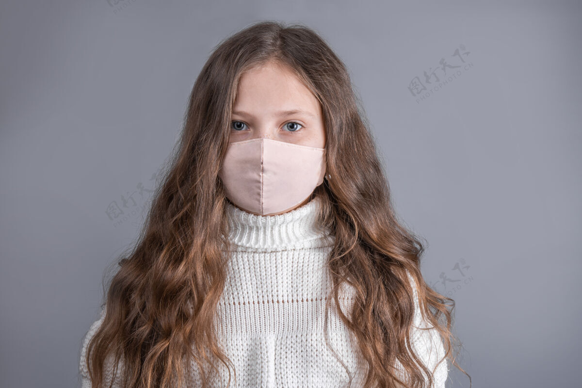 脸一个年轻漂亮的小女孩的肖像 金发飘逸 穿着白色毛衣 戴着医疗防护面具 在灰色的工作室里背景.地点为了文本.复制空间检疫疾病疾病