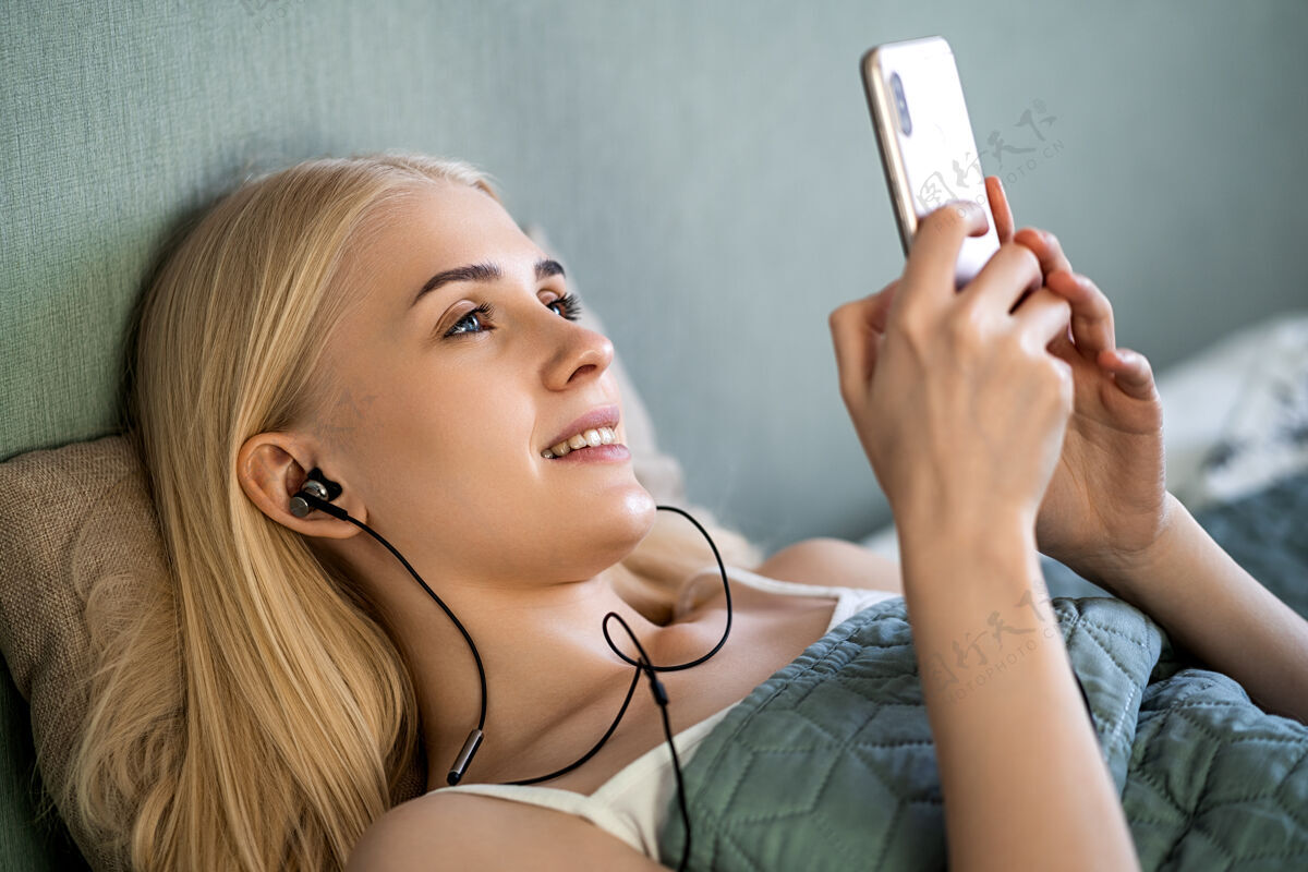 休闲微笑的女孩躺在卧室的床上用智能手机听音乐科技谎言床