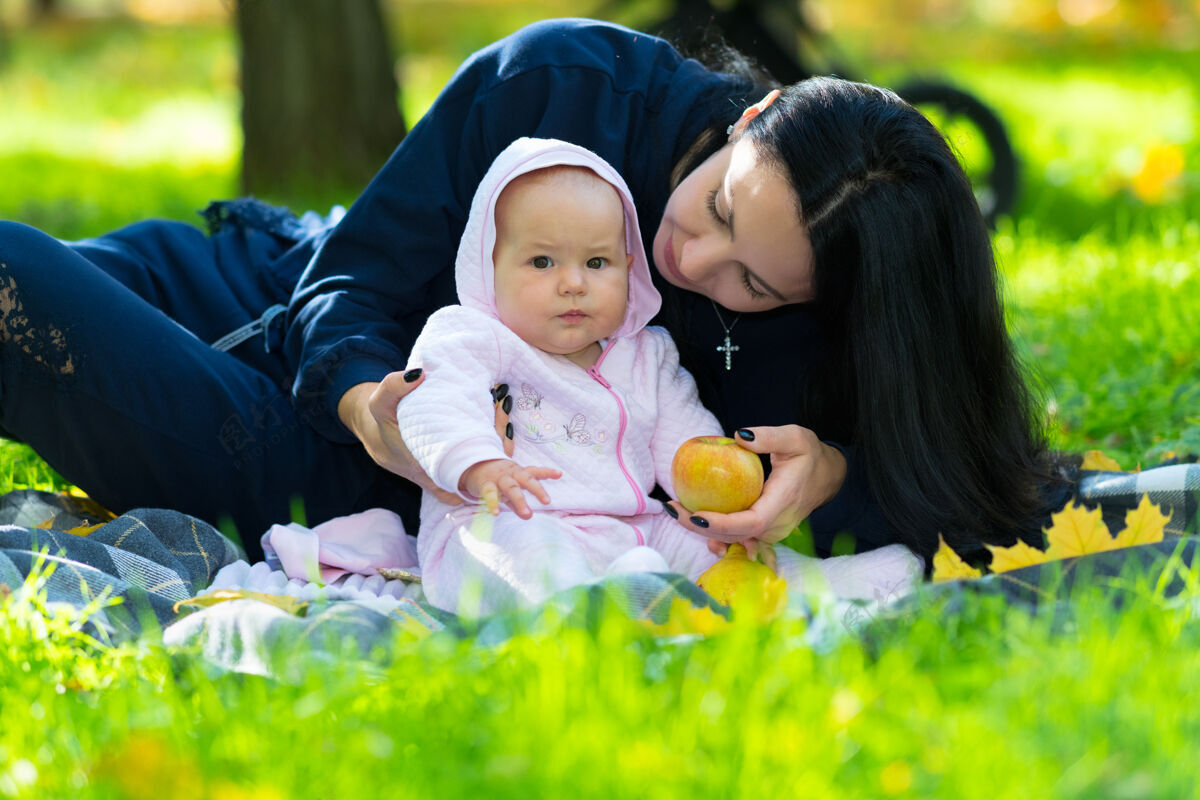 爱公园里 年轻的母亲带着她的小宝宝坐在绿草丛生的地毯上 给孩子一个秋天的苹果托儿举女人