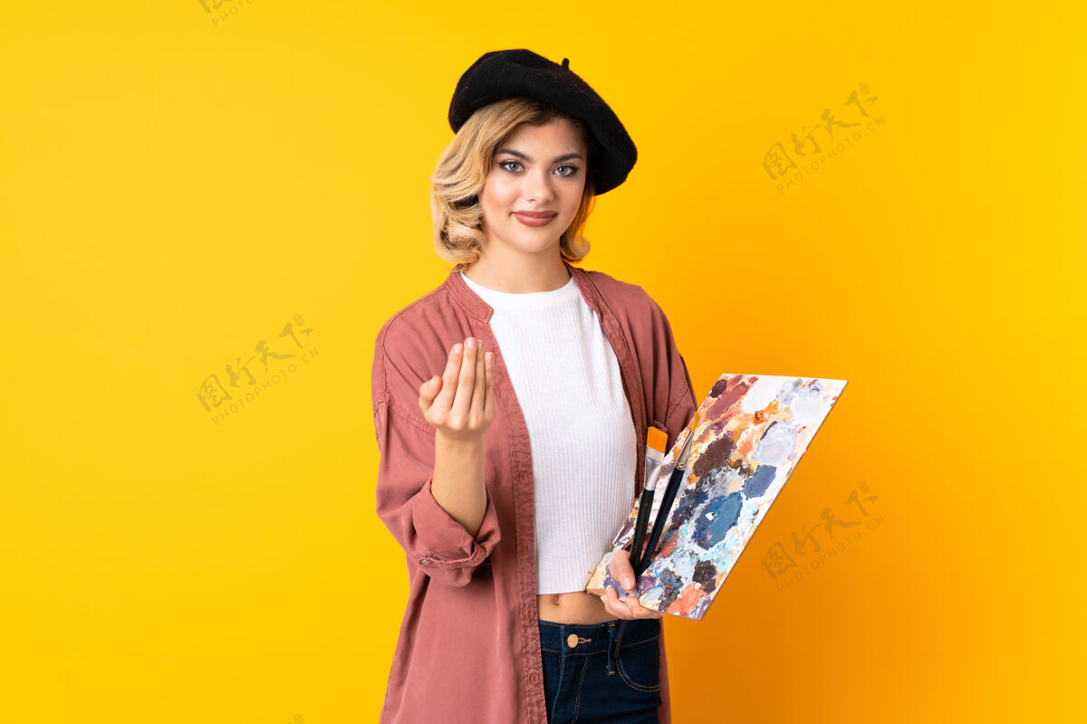金发年轻的艺术家女孩拿着一个调色板孤立在黄色邀请来俄罗斯展示绘画