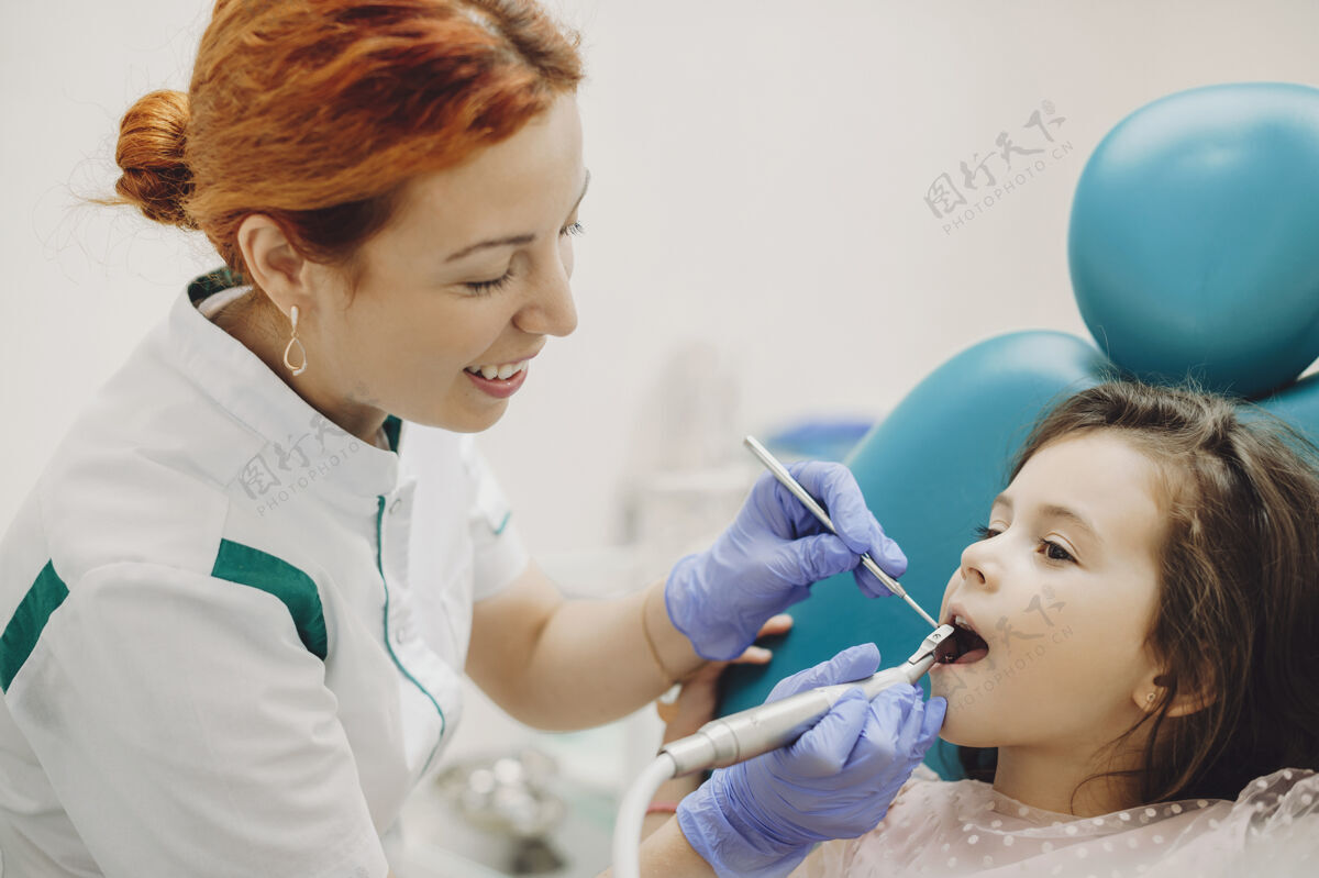 妇女一位年轻的女性儿童口腔医生为一个可爱的小女孩做牙齿手术的侧视图正畸专家椅子