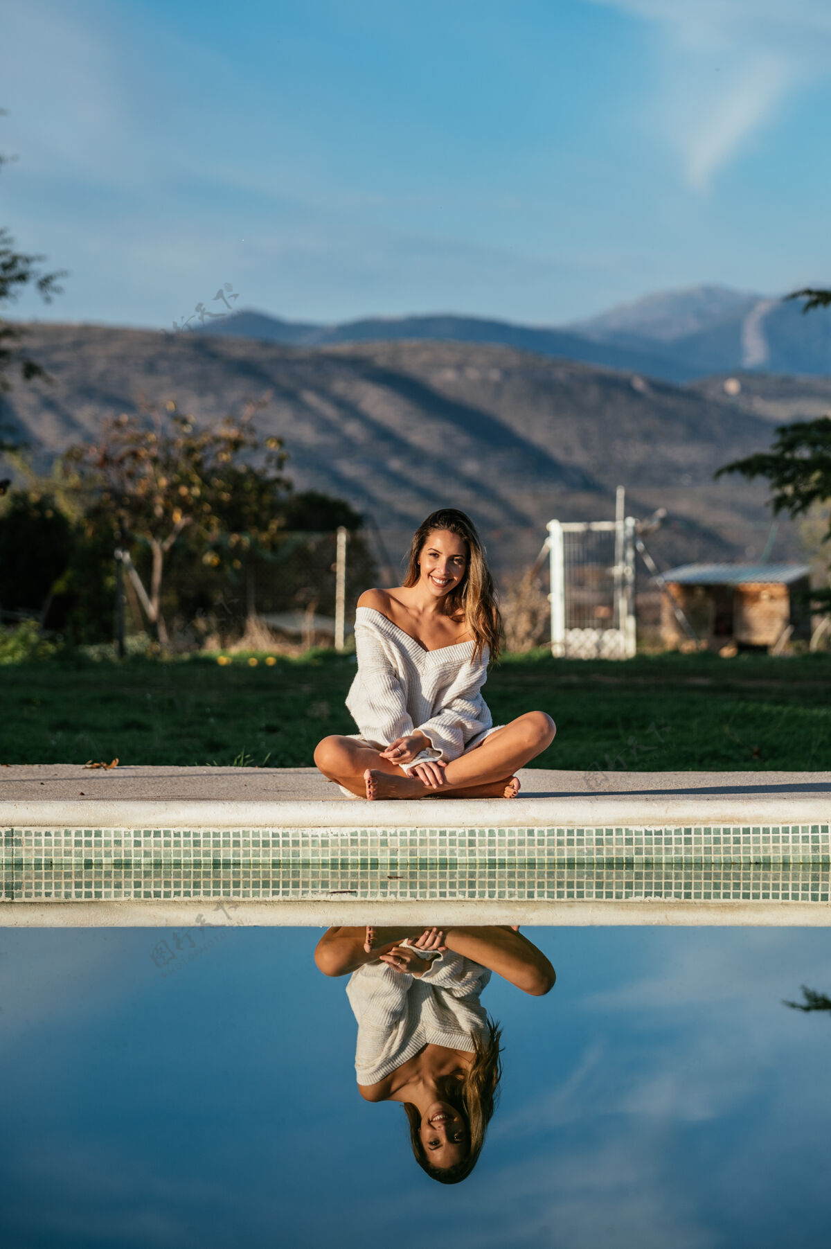 服装作物赤脚女性盘腿坐在游泳池附近与平静的反射水和微笑在院子里肖像漂亮休闲