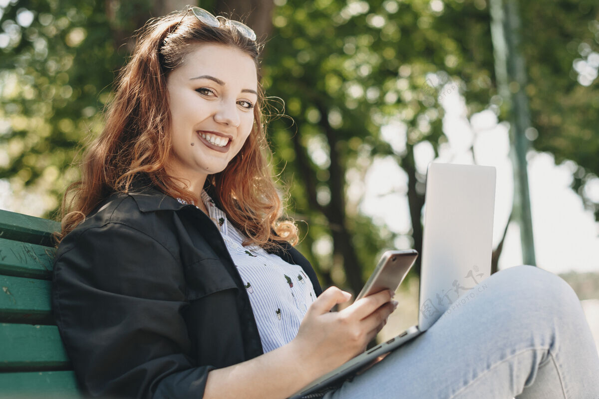 体重一个迷人的大个子女人坐在长凳上 腿上放着一台笔记本电脑 一只手拿着智能手机 微笑着看着相机脸微笑商务