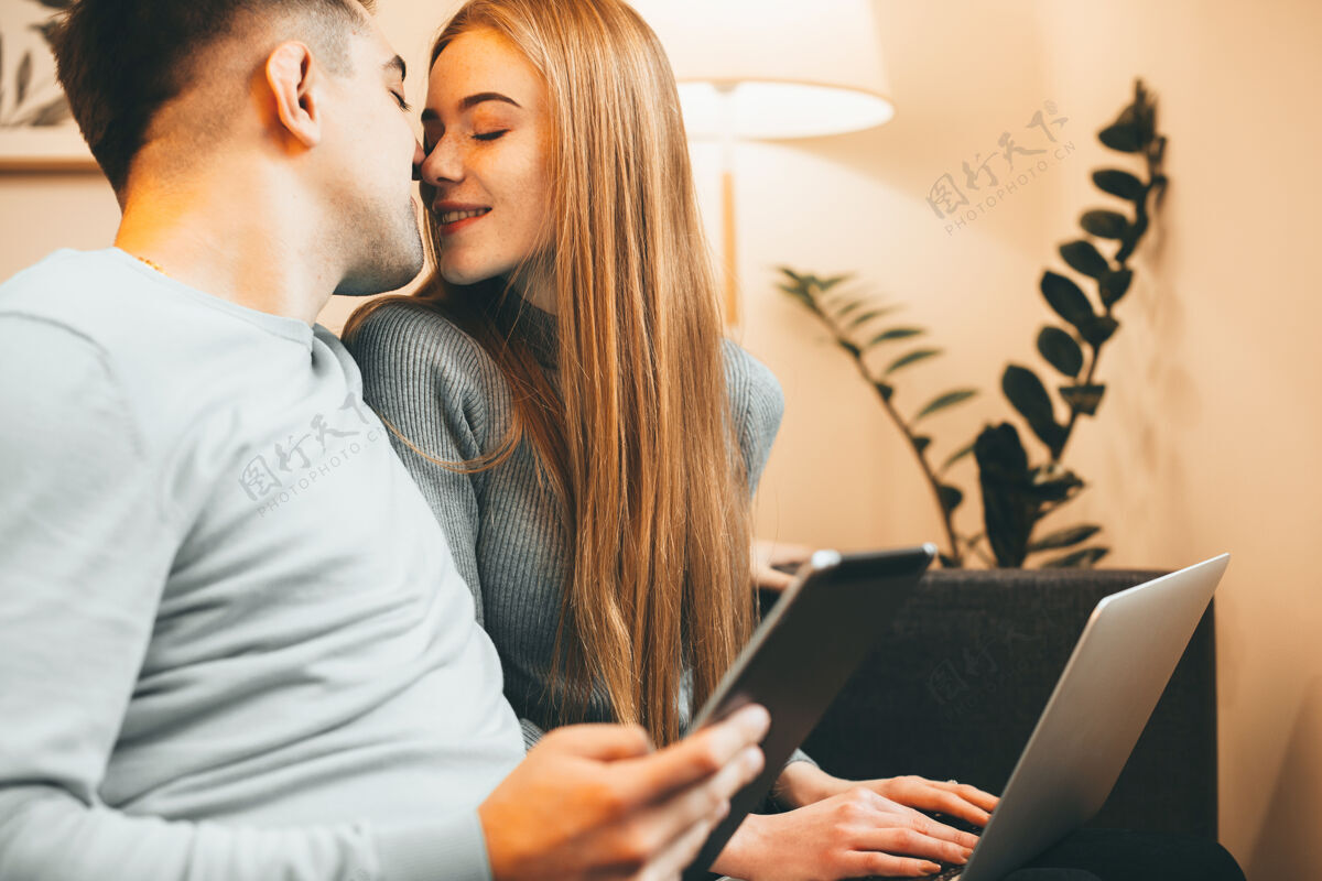 观看一对可爱的情侣坐在家里的沙发上 手拿笔记本电脑和平板电脑 微笑着亲吻的特写镜头帅哥手机微笑