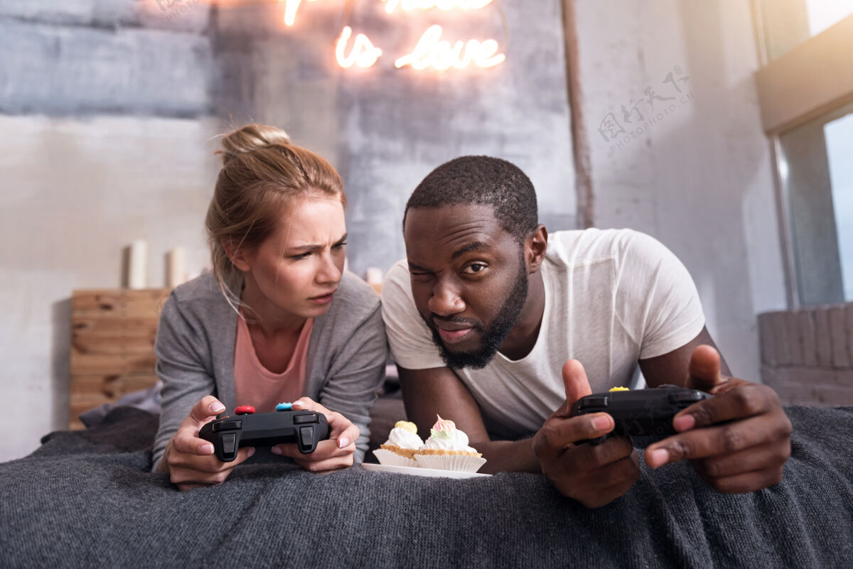 当代好笑游戏喜出望外高兴的国际夫妇吃纸杯蛋糕和玩电子游戏为什么享受周末爱情伙伴室内